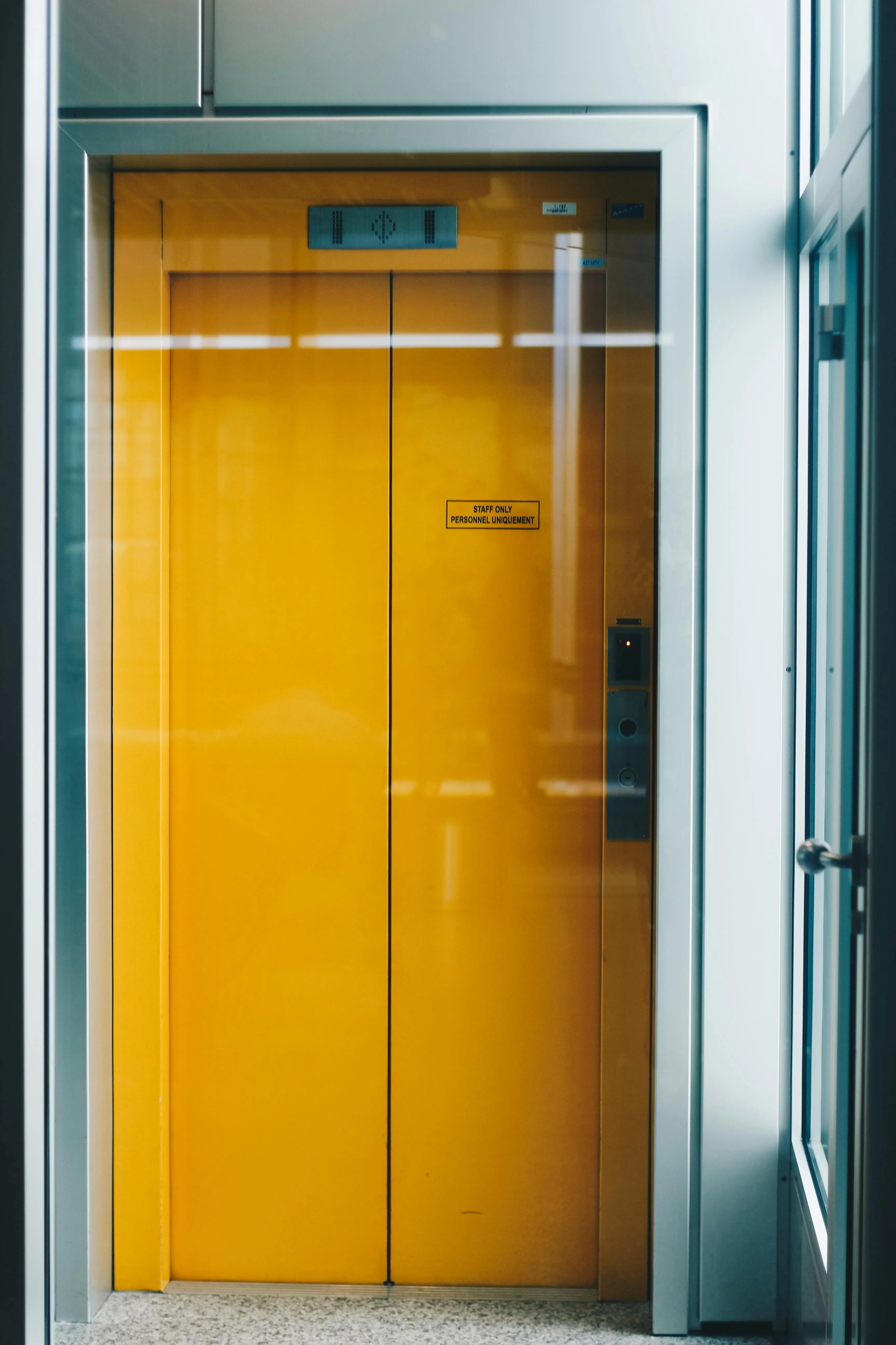 Porte d'ascenseur laquée jaune avec panneau de contrôle dans un couloir vitré